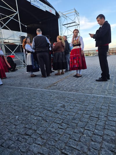 The image depicts a group of people in traditional Portuguese attire standing on a cobblestone surface, likely in Barcelos, Portugal. They are gathered near a stage structure. One man, dressed in a black suit with a red sash, is looking at a smartphone. The attire suggests a cultural event or performance.