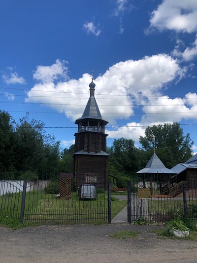 The image shows a wooden bell tower and adjacent structures of a religious nature, likely a church or monastery, situated in Izdeshkovo, Russia. The bell tower is a tall, dark wooden structure with a tiered roof topped with a small dome and cross. To the right, another building with a similarly shaped metal roof can be seen. A black wrought-iron fence encloses the property, with an open gate in the foreground. The grounds are grassy with scattered trees and some vegetation. A white trailer or small camper is parked on the grass within the fenced area. Power lines are visible crossing the sky, which is bright blue with scattered white clouds. The overall scene depicts a serene, rural religious site on a clear day.