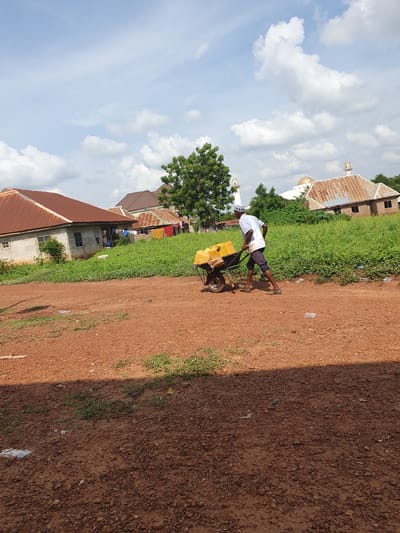 The image, originally rotated 90 degrees counter-clockwise, displays a daytime scene in what appears to be a rural or semi-urban area of Idah, Nigeria. The central subject is a man, dressed in a white short-sleeved shirt, dark shorts, and a white cap, actively pushing a yellow wheelbarrow. The wheelbarrow is loaded with several yellow jerrycans, suggesting he is transporting water or another liquid.
The man is situated on a reddish-brown dirt path or unpaved road, which occupies a significant portion of the foreground and midground. The path is uneven, covered with small stones and dirt, with patches of green grass and weeds growing along its sides. Sunlight casts shadows on the ground, indicating a bright, sunny day.
In the background, on either side of the path, various buildings are visible amidst green trees and vegetation. To the man's right, a structure with a golden dome and a white minaret, characteristic of a mosque, stands out. Another building with a white minaret is visible further to the left. Other houses with reddish-brown corrugated iron roofs and light-colored walls are scattered throughout the background. The sky above is clear blue with numerous white, fluffy clouds. There is no discernible text in the image.