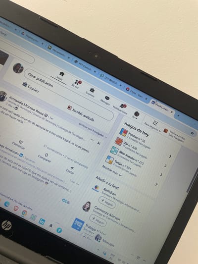 A medium-shot captures the upper portion of an open laptop, angled slightly towards the viewer. The screen displays the LinkedIn website, with a feed of articles, job postings, and game suggestions. The user interface language is Spanish. The laptop is situated on a light-colored surface, possibly a desk or table. The lower part of the laptop features the HP logo. The scene appears to be indoors, with natural light illuminating the screen. No people are visible in the frame. The city of Rionegro, Colombia is not identifiable from the visual information provided.