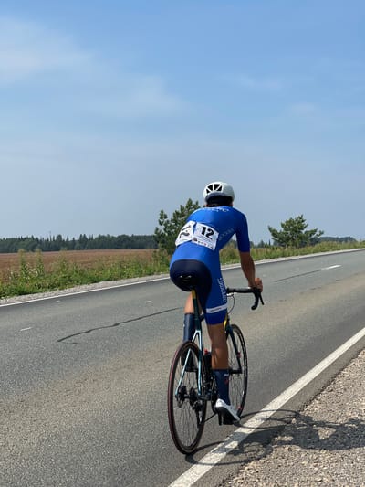 A lone cyclist wearing a blue and white racing suit and a white helmet is riding a bicycle on a paved road. The cyclist's bib number is 12, with the word "МОСКВА" visible on their legwear. The road has a white line marking the edge on the right. In the background, there is a field of brown vegetation bordering a line of trees. The sky is clear and blue with some faint clouds. The scene appears to be outdoors, possibly in a rural area. No specific landmarks or indicators point to the city of Niva, Russia.