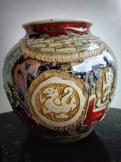 A close-up, eye-level shot shows an ornate, round ceramic vase with a rich glaze and raised decorative elements. The vase, with its wide opening and bulbous body, is positioned on a dark, reflective surface, likely a marble table, which casts a shadow. The background is a plain, light-colored wall.
The vase's exterior features a combination of textured and glazed surfaces. The rim is a deep, glossy red. Below the rim, a band of what appears to be brickwork is rendered in a pale, yellowish-tan glaze with darker grout lines. This is bordered by a fringe-like texture in a similar tan color. The body of the vase is adorned with intricate designs. A prominent circular medallion on the front displays a stylized creature, possibly a dragon or bird, in a lighter, matte finish against a textured gold background. To the right of the medallion, there are raised, angular symbols that resemble Chinese characters, rendered in the same light, matte finish against a gold textured background. The rest of the vase's surface is covered in a multi-colored glaze, exhibiting a swirling pattern of blues, purples, reds, oranges, and browns, creating a marbled or Raku-fired effect. This glazed surface reflects the surrounding environment, adding depth and complexity to the image.
The location of Kualaraja, Indonesia is not visible in this image.