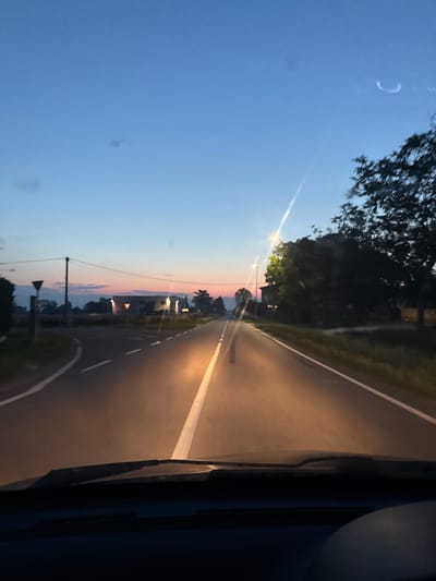 The image depicts a view from inside a vehicle on a rural road at dusk. The sky transitions from a deep blue at the top to hues of pink and orange near the horizon. A road stretches ahead, illuminated by the headlights of the vehicle, with a white line marking the center. To the right, trees and vegetation line the road, and a street lamp is visible. On the left, a road diverges, with a yield sign and a triangular traffic sign. In the distance, buildings are visible, including one with illuminated windows. The scene is consistent with a rural setting in the vicinity of Villa Pasquali, Italy.