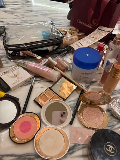 An assortment of makeup and skincare products is scattered on a marble surface. Notable items include Charlotte Tilbury products such as a burgundy tote bag, blush compacts, and an Airbrush Flawless setting spray. A blue Medicube Zero Pore Pad container is visible. Other makeup items consist of eyeshadow palettes, foundation, concealer, brushes, and a clear travel makeup bag containing miscellaneous items. A Chanel compact is also present.