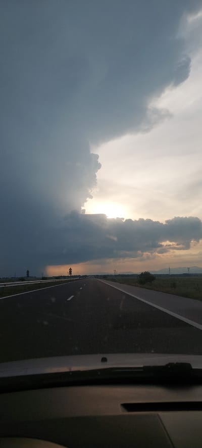 The image depicts a rural highway scene at what appears to be late afternoon or early evening, likely near Palauzovo, Bulgaria. The perspective is from inside a vehicle, with part of the dark interior visible on the far right.

A two-lane asphalt road with white solid and dashed markings extends into the distance on the right side of the frame. Beyond the road, the landscape is flat and green, with distant hills or mountains faintly visible under a hazy sky. Several utility poles and power lines are scattered across the distant terrain, and a few small trees dot the landscape. Two dark, indistinct road signs on poles are present on the roadside in the middle distance.

The left side of the image is dominated by a dramatic sky. Heavy, dark grey storm clouds gather on the far left. Towards the center, brilliant sunlight breaks through lighter, textured clouds, creating a bright, almost ethereal glow. This contrast between the dark, imposing clouds and the radiant light suggests dynamic weather conditions, possibly a passing storm or the lead-up to sunset. The overall mood is one of awe due to the powerful natural elements. No visible text is present.
