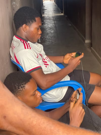 Two young black men lie on the ground under a dark structure, possibly a bridge or overpass. One wears a white and red soccer jersey, the other a white t-shirt and dark shorts. They sit in blue plastic chairs, appearing cramped and close together. Each holds a smartphone and seems to be engrossed in playing games. A charging cable dangles between them. The scene suggests a makeshift gaming spot in an unusual or less than ideal location.