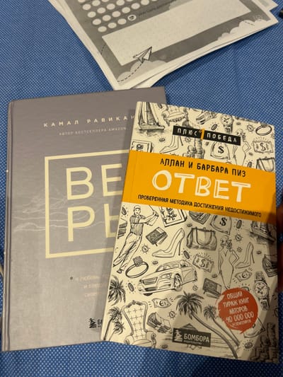 This image shows a top-down view of two books and some papers on a blue, patterned surface. The top book is a bright yellow and white cover with a black and white drawing of various symbols of wealth and luxury, including jewelry, money bags, a sports car, and high heels. The title of the book is in large, bold orange letters, "OTBET," and smaller text above and below it reads "АЛЛАН И БАРБАРА ПИЗ" (Allan and Barbara Piz) and "ПРОВЕРЕННАЯ МЕТОДИКА ДОСТИЖЕНИЯ НЕДОСТИЖИМОГО" (Proven Method for Achieving the Unachievable). There is also a red circular sticker with text in Russian. The second book, lying partially underneath the first, has a dark grey cover with white text. The visible text on this cover includes "КАМАЛ РАВИКАН" (Kamal Ravikant) and "АВТОР БЕСТСЕЛЛЕРА АМАZON" (Author of Amazon Bestseller). Large white letters on this book spell out "BE P". To the left of the books are some papers with white circles and dotted lines, possibly from a planner or notebook, and some Russian text. The scene appears to be indoors, with soft lighting, and the overall impression is one of study or leisure. The specific location context is Sabunchu Raion, Azerbaijan.