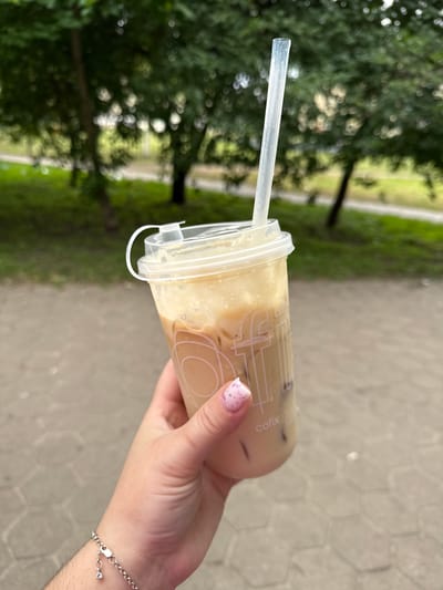 A hand with a silver bracelet and pink speckled nail polish holds a clear plastic cup of iced coffee with a white straw. The cup has a logo that reads "cofix". The background shows trees and a paved walkway. The image was taken in Stary Oskol, Russia.