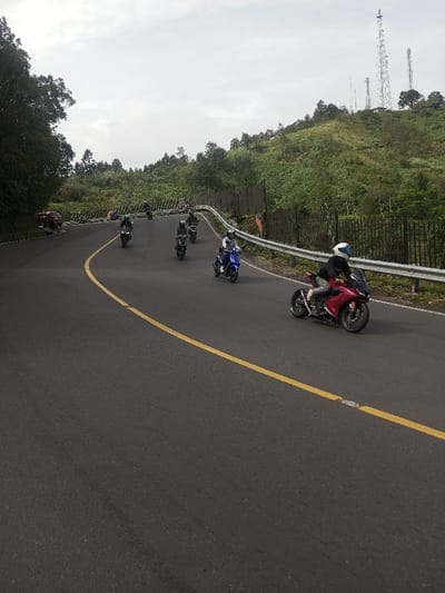 A group of motorcyclists travel on a winding paved road, marked with a yellow line. Riders wear helmets and a mix of light and dark clothing. A red motorcycle is closest to the camera, with other motorcycles following behind. A guardrail runs along the road's edge, and trees and communication towers are visible on the hillside in the background. The sky is overcast.