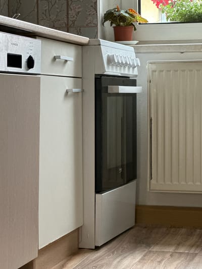 A slightly high-angle, medium shot captures a domestic kitchen corner in Bad Blankenburg, Germany. The scene is dominated by a white electric oven with a glass door, positioned next to a beige cabinet with two drawers. The oven has a control panel with multiple circular knobs. To the left of the oven is a section of a light wood-grain cabinet with a silver handle and a built-in control panel labeled "VORWERK" with a black display and a dial. The countertop is a light wood-grain finish, with a subtle floral wallpaper in the background. A window above the oven reveals a potted plant with red and green foliage. A cream-colored radiator is visible to the right of the oven. The floor is covered with light brown wood-grain laminate flooring. The lighting suggests an indoor setting with natural light coming from the window.