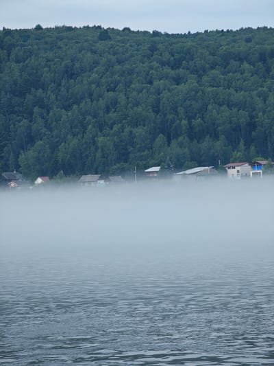 The image depicts a serene, misty landscape in the village of Atamanovo, Russia. In the foreground, a body of water, likely a river or lake, displays gentle ripples under a hazy sky. A dense, green forest blankets a hillside in the background. Nestled at the base of the hill, partially obscured by thick fog, is a small settlement of houses. The houses appear to be of various sizes and styles, with some featuring visible roofs and windows. The overall atmosphere is tranquil and somewhat mysterious due to the pervasive mist. No people or animals are visible in the scene.