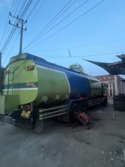 In Matangpanyang, Indonesia, an outdoor scene depicts a light green and blue tanker truck parked on a dirt or gravel lot. The truck is the primary subject, occupying the center and right side of the frame. A person, wearing an orange garment, is crouched beside the truck's rear passenger-side wheels. The truck has a ladder on its rear, and a license plate is visible. In the background, a partially constructed building with columns and a roof is present. Overhead, power lines stretch across the sky, with a utility pole to the left. The sky is a clear, pale blue, suggesting either early morning or late evening. A stack of tires is visible to the far right of the image.