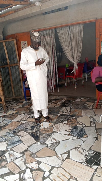The image depicts a man standing in what appears to be a covered, open-front establishment in Dakata, Nigeria. The man, with a dark beard, wears a white embroidered tunic over white trousers and dark sandals, topped with an embroidered skullcap. He holds a silver-colored smartphone with both hands, looking down at it. The floor is covered with an irregular mosaic pattern composed of various broken ceramic tiles in shades of white, beige, brown, grey, and black. To the man's left, corrugated metal sheets are visible, along with a blue plastic chair. To his right, an entrance framed by orange-painted wood leads into an inner room. A light-colored string curtain hangs in this doorway, partially tied back. Inside the room, several red plastic chairs with metal frames are arranged around a table, on which a green mug is visible. A black electric fan is mounted on the far wall of the inner room. A partial view of a woman in a pink top is visible on the far right, seated on a red chair. The ceiling is rough concrete with visible wooden support beams and an exposed white LED bulb.