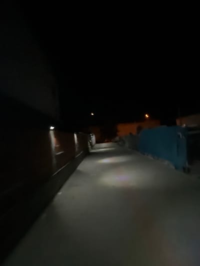 The image depicts a nighttime scene in the city of Novi Iskar, Bulgaria. The perspective suggests a street or alleyway. On the left side of the frame, a dark, imposing structure, possibly a building wall, occupies a significant portion of the foreground. To the right, a narrow pathway or road stretches into the darkness, illuminated by faint light sources. The pathway is bordered by what appear to be low walls or fences. Further down the path, a bright blue structure is visible, suggesting it may be a large dumpster or a storage container. A faint orange light is visible in the distance, possibly from a street lamp or a window. The overall atmosphere is dimly lit and somewhat desolate. No people are visible in the image, and no specific actions or interactions are captured. The environment appears to be urban, characterized by concrete or asphalt surfaces and artificial lighting.