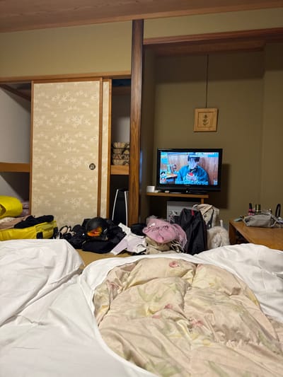 This image captures the interior of a room in Hakuba, Japan, from a low perspective, likely from a floor-level bed. In the immediate foreground, two futon-style beds are visible, both covered with white sheets. One of the futons features a light-colored duvet with a floral pattern.

The room, characterized by olive-green walls and dark wood structural elements including a central pillar and ceiling beams, shows signs of recent occupancy. To the left, a yellow duffel bag, several black bags, multiple pairs of shoes, and a black ski helmet with an orange-tinted visor are arranged on the floor near a set of light-patterned sliding doors. A white PlayStation 5 console stands upright beside the central wooden pillar. The sliding doors are partially open, revealing shelving with folded bedding or cushions.

In the midground, a black flat-screen television is displaying a Japanese program featuring two male figures, one holding a red object. The TV rests on a low wooden stand, beneath which a white rectangular appliance is positioned. To the right of the television, various items of clothing, including a pink scarf and a tote bag with a "Paris" motif, are piled on the floor. A small, fluffy white dog is also visible, resting on the floor. Further to the right, a low wooden table or bench holds several bottles and a grey bag. Above the television, a small, framed piece of artwork depicting a stylized plant hangs on the wall. No individuals are directly visible within the room, only on the television screen.