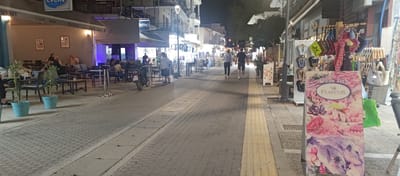 This image depicts a pedestrianized commercial street at night within the Municipal Unit of Ellomenos, Greece. The street is paved with light-colored interlocking bricks, featuring a central pathway and sidewalks on either side. A prominent yellow tactile paving strip runs along the right sidewalk.

On the left side of the street, an establishment named "EVENT CAFE BAR" is visible, featuring an awning over an outdoor seating area. Multiple individuals, approximately eight to ten, are seated at various tables and chairs, engaged in conversation or dining. Two potted plants in turquoise pots are placed on the ground in front of the seating area. Further down the street on the left, another business has a sign displaying "CENTRO" and an "HAPPY HOUR" offer for cocktails at 5.00 €. A black motorcycle with a Greek license plate "EYK 7212" is parked near the left sidewalk, adjacent to the outdoor seating of "EVENT CAFE BAR". The buildings on the left are multi-story with balconies, characteristic of local architecture.

The right side of the street features shopfronts with merchandise displays extending onto the sidewalk. In the immediate foreground, an upright rectangular sign advertises "Fresh fish FLISVOS" with colorful illustrations of seafood including fish, octopus, lobster, shrimp, and mussels. Adjacent to this sign, racks of various items such as handbags, colorful scarves or beachwear, and flip-flops (some bearing the "ILSE JACOBSEN" brand) are displayed. Further along the right, a traditional-style streetlight illuminates the area.

In the center of the street, two pedestrians, a woman in light clothing and a man in a dark t-shirt and shorts, are walking away from the camera. The street is well-lit by artificial lights emanating from the businesses and streetlights, creating an illuminated evening atmosphere. Shadows are cast by people and objects due to the varied light sources.