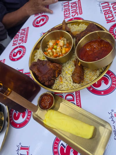 This is a top-down shot of a serving of Middle Eastern cuisine, likely Kabuli Pulao, presented on a brass platter. The dish consists of rice, pieces of meat (appearing to be lamb or chicken), and two small bowls containing sauces or relishes. One bowl contains a vibrant red, chunky sauce, while the other holds a finely diced salad of vegetables like cucumbers, carrots, and possibly onions. To the right of the platter, a golden tray holds a long, pale yellow item, possibly a type of bread or a sweet confection.
The food is laid out on a white surface that has a repeating pattern of red and white circular logos with the letter "S" inside. Text in both Arabic and English is visible on this surface, including "KEBULI SAUDI," "AYAM & KAMBING" (which translates to "Chicken & Goat" in Indonesian), and Arabic script that appears to read "كبولي سعودي" (Kabuli Saudi). This branding strongly suggests the restaurant is "Kebuli Saudi" and specializes in dishes like Kabuli Pulao, possibly offering chicken and goat variations.
A wooden utensil with a metal tip, likely for serving or eating, rests near the golden tray. In the lower left corner, a person's left arm and hand are partially visible, dressed in dark clothing. The overall lighting suggests an indoor setting, possibly a restaurant, with no clear indication of the time of day or weather. The focus is entirely on the food presentation and branding.