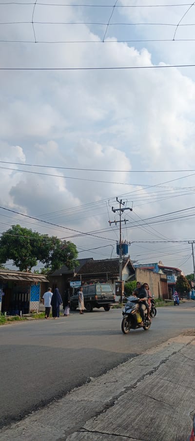 The image depicts a street scene in Sukarame, Indonesia. In the foreground, the asphalt road shows signs of wear and tear with cracked concrete edges. A black motorcycle with two riders is moving away from the viewer. The female rider, wearing a dark hijab, is driving, and a child is seated behind her. Further down the road, a dark green SUV is parked.
On the left side of the street, a small blue and white structure, possibly a shop or kiosk, stands with several bottles visible inside. A group of four people, dressed in traditional Islamic attire, are standing near this structure and the road. Two individuals are facing away from the viewer, while two others are looking towards the parked SUV.
In the background, several houses with dark tiled roofs are visible. A prominent utility pole with transformers and numerous power lines stretches across the sky, which is filled with scattered white clouds against a pale blue backdrop. Trees with dense green foliage are also present. To the right, another motorcycle ridden by a person in a pink headscarf is visible in the distance. The overall lighting suggests it is late afternoon or early morning.