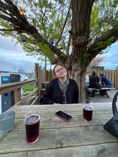 A vertical, full shot shows a woman at a wooden table, with a glass of dark liquid, possibly beer or wine, and a cell phone on the table. The scene is outdoors, with a large tree to the left decorated with multi-colored lights. In the background, other people are sitting at a table. A fence and a building are visible beyond the tables. The building displays the name "PADRE JOAK" and indicates it is for electric vehicle charging. "HOTHER" is also on the building. The location is Villeneuve-d'Ascq, France.