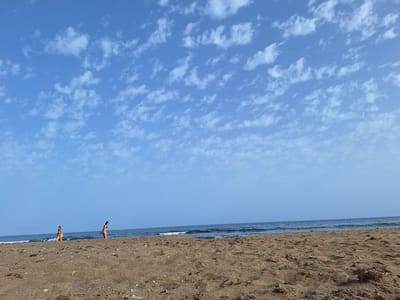 A wide shot captures a tranquil beach scene with two individuals present. The foreground is dominated by a sandy beach, sparsely littered with seaweed and small shells. In the middle ground, two women, dressed in swimwear, are walking near the water's edge. The sea is calm with small waves rolling onto the shore. The horizon is a clear blue, meeting a sky filled with scattered white clouds. The image appears to be taken from a low vantage point on the beach, looking out towards the ocean.