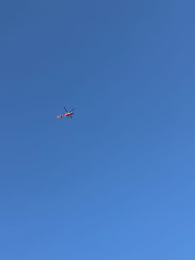 A red helicopter with black rotors and a black tail rotor is captured in flight against a clear blue sky. The helicopter is positioned in the upper left quadrant of the image. The background is entirely composed of the vast expanse of the sky, with no visible ground features, buildings, or other objects.