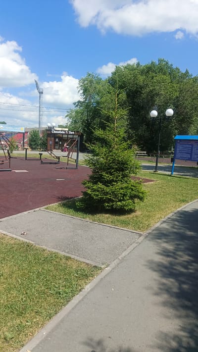 A wide shot captures an outdoor scene in the city of Kuybyshev, Russia, on a sunny day with scattered white clouds in a blue sky. In the foreground, a small evergreen tree stands on a patch of grass bordered by a paved walkway. To the left, a reddish-brown rubberized surface, likely part of a playground or sports area, is visible, featuring outdoor exercise equipment. A woman is seen inside a small building with a sign that reads "Chocolate Box." In the background, a tall stadium light tower and dense green trees are prominent. Further to the right, a lamppost with two globe lights stands near a blue informational sign. The scene is peaceful and appears to be in a public park or recreational area.