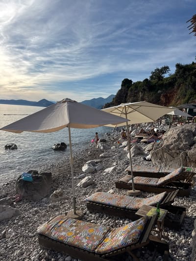 The image depicts a rocky beach in Rijeka Reževići, Montenegro, under a partly cloudy sky. Several beach chairs with colorful cushions are arranged on the pebble-strewn shore, some occupied by people sunbathing or relaxing. Two large, light-colored umbrellas provide shade. The calm sea stretches out towards a hazy horizon with mountains in the distance. A child with a pink toy is visible near the water's edge, and a green bottle sits on a small table next to one of the lounge chairs. The environment is natural, with rocky cliffs and vegetation visible on the right side of the frame.