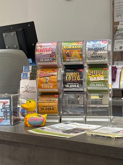 A close-up, eye-level shot shows a display of lottery tickets and a decorative yellow duck on a grey counter, likely in a retail setting in Hinterzarten, Germany. The lottery tickets are arranged in clear plastic holders, with different denominations and prize amounts visible. Some of the ticket names include "WILD CASH," "EURADO," "DIAMANT," "Verrückte 7," "RUBBEL LOTTO," and "SUPER CASH." A yellow duck figurine, decorated with colorful patterns, sits in front of some of the ticket displays. The background is slightly blurred, showing a wall and possibly shelves with more items. The overall scene suggests a point of sale in a shop where lottery tickets are sold.