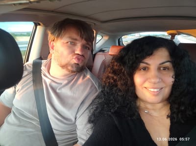Two individuals, a man and a woman, are in the front seats of a car. The man, on the left, is facing the camera with his lips pursed, making a duck face. He has short brown hair and is wearing a light grey t-shirt and a seatbelt. The woman, on the right, is smiling at the camera. She has long, curly dark hair and is wearing a black top and a delicate necklace. The car's interior is visible, including leather seats and a sunroof. Outside the car windows, a landscape with fields and a road is visible, suggesting they are traveling. The timestamp "13.06.2026 05:57" is superimposed on the image. The location is stated to be Shtarkovo, Bulgaria.