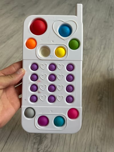 A white, toy cell phone-shaped pop-it toy is lying on a light wood-grained floor. The toy has several colored silicone bubbles. At the top, there's a red bubble, then an orange bubble below it. To the right of the orange bubble is a clear bubble, and to the right of that is a yellow bubble. Above the yellow bubble and to the right of the red one is a blue bubble inside a heart-shaped indent. To the right of the blue bubble is a green bubble. Below these top bubbles is a keypad area with rows of purple bubbles and white heart shapes. The bottom of the toy has three more bubbles: grey on the left, purple in the middle, and teal on the right. A person's hand is visible on the left side of the toy, holding it up slightly. The background is the floor. There is no information provided about the location in the city of Мариинский, Russia, in this image.