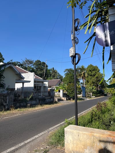 The image depicts a quiet street in Pagowan, Indonesia, on a clear, sunny day. The road, paved with asphalt and marked with a white line, is bordered by a grassy verge and a concrete barrier in the foreground. On the left side of the road, there is a low, light-colored house with a tiled roof, a fence, and some vegetation. Beyond the house, a line of trees separates the residential area from the sky. On the right side of the road, a utility pole stands tall, carrying numerous black cables and a small grey box labeled "PJM 200-30." Adjacent to the pole, a hanging sign, possibly for a business named "Nice," is visible, along with the trailing fronds of a dragon fruit plant. Further down the road, more trees and houses can be seen, with a blue gate visible in the distance. The overall impression is one of a peaceful, rural or suburban environment in Indonesia.