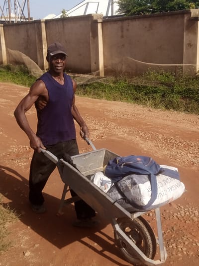 A man in a dark blue sleeveless sweater and black pants pushes a wheelbarrow filled with two large bags and a blue backpack. The man wears a dark baseball cap. The wheelbarrow is on a dirt road. In the background, a light brown wall with pillars separates the road from an area with green grass and netting. Trees and buildings are visible beyond the wall. The location is Katon Rikkos, Nigeria.