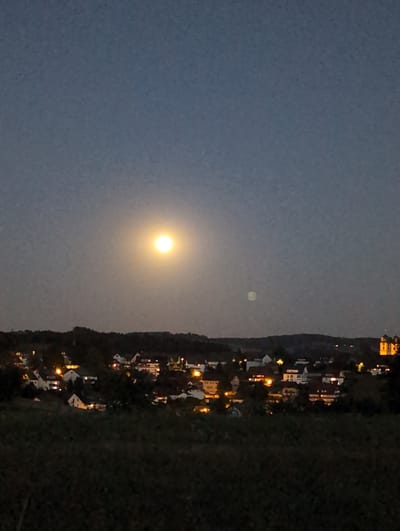 The image shows a nighttime scene of a village with the moon visible in the sky. The moon is bright and appears to be full, casting a yellowish light. The village is situated on a gentle slope, with houses and buildings illuminated by lights. The architecture suggests a European setting, possibly in the region of St. Peter, Germany, given the characteristic steep roofs and the presence of a church spire visible in the distance on the right side of the image. The foreground is dark, appearing to be a field or vegetation. The sky is a deep blue, characteristic of twilight or early night. There are no people or animals visible in the image. The overall atmosphere is serene and peaceful.