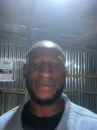 This is a close-up, portrait-style photograph of a Black man taken from a low angle, with the camera tilted. He appears to be indoors, in a dimly lit space with corrugated metal walls and ceiling, suggesting a rustic or informal setting. The lighting is dramatic, with a strong light source to the left of the frame creating bright highlights on the subject's face and the metal surfaces.
The man has short hair and a beard, and he is wearing a light blue denim jacket over a dark shirt. His expression is neutral, and he is looking directly at the camera. His face is partially illuminated, with some areas in shadow.
In the background, a sign is visible with text that appears to be related to a snooker club or arcade. The visible text includes "PLUG PLAY HUB", "SNOOKER ACCOUNT", "8105536869", "OPAY", "1 GAME N300", "2 GAMES N500", and "EXPRESS N500". This suggests the location is a place where people can play snooker and other games, and it appears to be a business in Nigeria, given the currency notation "N" for Naira. The time of day is not explicitly indicated, but the artificial lighting and dim environment suggest it could be evening or night.