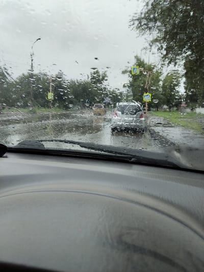 The image depicts a view from inside a vehicle looking out through a rain-streaked windshield onto a flooded street in Chernogorsk, Russia. It appears to be a grey, overcast day with heavy rain. Several cars are visible on the road, some appearing to be stationary or moving slowly through the water. Traffic signs, including a pedestrian crossing sign, are visible on poles along the roadside. Trees line the street, their foliage dark and wet. The foreground shows the interior of the car, including the dashboard and part of the steering wheel. The scene conveys a sense of difficult driving conditions due to the rain and flooding.