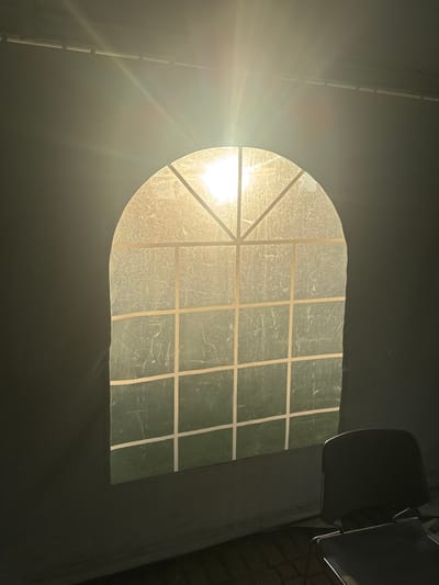 The scene depicts an interior space in Waltham, United States, illuminated by intense natural light emanating from a centrally positioned arched window. The window is segmented by a grid pattern and appears to be composed of a translucent, textured material that obscures the view beyond, while allowing direct sunlight to penetrate. The sun's position is visible through the upper portion of the window, producing a bright, yellowish-white glow and prominent lens flares. The lower part of the window surface exhibits a greenish hue, suggesting a verdant exterior. The surrounding interior wall is dark, appearing to be a flexible or fabric-like material, possibly part of a tent or temporary structure, with a visible horizontal structural element at the top of the frame. In the lower right foreground, a dark, unoccupied stacking chair is partially visible, primarily in shadow, resting on a dark, patterned floor. No individuals are present in the image.