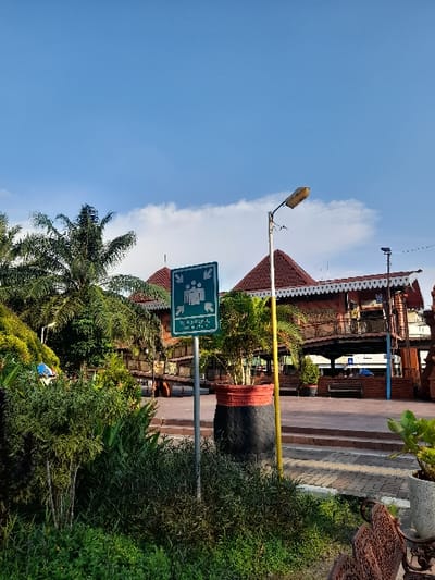 The scene is located at -7.464 latitude and 112.432 longitude in Mojokerto, Indonesia. A green sign with the word "TURUN KEREPA" and an image of people walking in different directions is attached to a wooden pole in front of a red-roofed building. The building appears to be a station or a terminal with multiple entry/exit points, including a shaded walkway with an open-air area and a brick platform. Green bushes are in front of the building and on both sides of the wooden pole. A streetlight is seen on the right side of the pole, and a potted tree is in front of the building. A couple of benches are on the ground in the foreground of the image. The sky is blue and cloudless.
