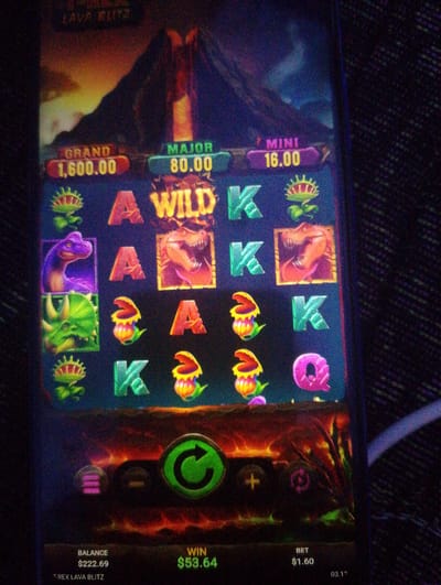 The image displays a screenshot of a mobile slot machine game titled "T-Rex Lava Blitz." The game features a volcanic eruption background and various symbols on the reels, including dinosaurs, a Venus flytrap, and card game letters (A, K, Q). Jackpot amounts labeled "GRAND," "MAJOR," and "MINI" are visible, with current values of $1,600.00, $80.00, and $16.00 respectively. At the bottom of the screen, the player's balance is $222.69, the current win is $53.64, and the bet is $1.60. A large green circular button, likely for spinning the reels, is centrally located at the bottom. The game appears to be running on a mobile device held by someone, as evidenced by the slightly distorted image and the dark, textured background which suggests the device is in a low-light environment. There is no discernible information within the image to indicate any specific city or location, including Pasirdoton, Indonesia.