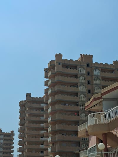 The image shows several tan-colored apartment buildings with a distinctive castle-like design, featuring crenellated tops and arched windows with decorative grates. The buildings are tall and appear to be located in a residential area. In the foreground, a section of a building with balconies and an outdoor staircase is visible. The sky is a clear, light blue, suggesting a sunny day. There is no visible signage or explicit indication of the location within the image itself, but these buildings are known to be the "Castillo de Don Juan" apartments in the city of San Javier, Spain. There are no people or animals visible in the frame.