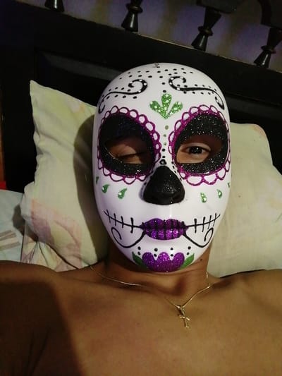A person wearing a white sugar skull mask lies against a pillow, facing the camera. The mask is decorated with black, green, and purple glitter detailing. The eye holes are lined with black glitter, and the mouth is purple glitter. The person wears a gold necklace with a cross pendant. A bed's headboard is visible in the background.