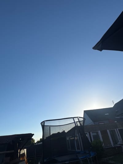 This residential backyard scene, stated to be in Pirmasens, Germany, depicts a bright, clear blue sky with the sun visible as a distinct glow on the lower right horizon. The image is significantly backlit, casting foreground and midground objects into silhouette. A large, dark-colored trampoline with a safety net enclosure occupies the midground, its structure clearly defined against the lighter sky. In the upper right corner, a segment of a dark patio umbrella is visible, extending into the frame.
On the far left, a dark-roofed shed or outbuilding with exposed wooden framing is partially visible, also silhouetted. In the background, a multi-story house with a dark, steeply pitched tiled roof is prominently positioned on the right, partially obscuring the sun. Above this house, utility wires and a support structure are visible. Adjacent to and in front of this main house, a single-story extension or sunroom with a corrugated roof and multiple vertical window panes is discernible, situated behind the trampoline. Minimal vegetation, including some trees and bushes, is scattered throughout the scene, primarily in silhouette. No people are present.