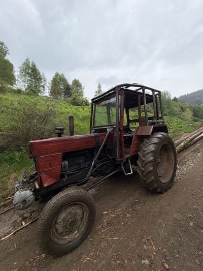 A red tractor with a roll cage is parked on a dirt road at an angle. The front tires are smaller than the rear tires. Both tires are dirty. The tractor is parked near green vegetation and a hillside. The sky is overcast. The location is in Rilski Manastir, Bulgaria.