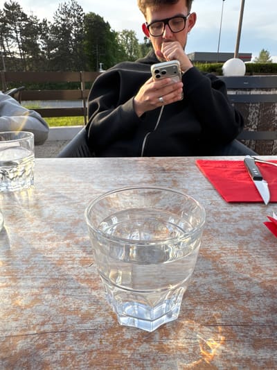 Here is a forensic description of the image:

The image shows a person seated at a table outdoors. The person is holding a smartphone and appears to be taking a photograph. On the table, there is a partially-filled clear glass of water, a red napkin, and a butter knife. The table surface has a weathered wood grain. In the background, there are trees and a cloudy sky. Based on the provided geo-location, the scene is located in Les Ulis, France. The image is rotated counterclockwise.