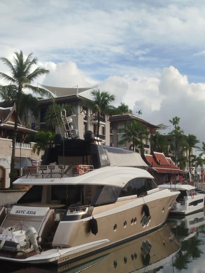 A large, beige yacht named "BAYC ASIA" is docked in a marina. The yacht is in the foreground, with its stern facing the viewer. A smaller white boat with an outboard motor is attached to the stern of the larger yacht. In the background, several buildings with distinctive Thai architecture, featuring ornate roofs and balconies, are visible. Palm trees are scattered throughout the scene, and the sky is partly cloudy. The image appears to be taken from a slightly elevated position, looking down at the yacht and the marina. The location is identified as Ko Kaeo, Thailand.