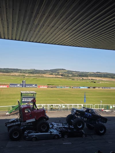 The image captures a sunny outdoor scene at Punchestown Racecourse, Ireland, likely during an event involving monster trucks. In the foreground, a red monster truck with large tires is positioned over several crushed cars. A person in an orange outfit stands on top of the red monster truck, appearing to wave or pose. To the right, a black monster truck is also positioned over crushed cars. The background shows a vast green field, typical of a racecourse, with distant hills and trees under a clear blue sky. Several banners and signs are visible in the mid-ground, including "PUNCHESTOWN" branding and advertisements. The overall atmosphere suggests a lively event with a focus on automotive stunts.