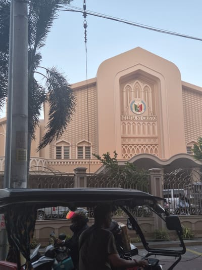 A daytime, eye-level shot shows the Iglesia Ni Cristo (Church of Christ) building in Pasay, Philippines, partially obstructed by a passing vehicle, likely a tricycle, on the right. The cream-colored building has a pointed arch facade with "Iglesia Ni Cristo" inscribed vertically. A circular emblem sits above the inscription. The building is textured with patterns. Three windows are visible on the lower portion of the building. A metal grate fence adjoins the building. A utility pole and palm fronds are visible on the left side of the frame. Power lines stretch across the upper portion of the image. The sky is pale blue.