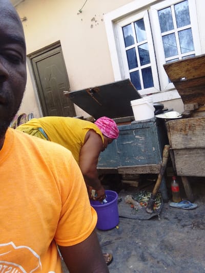 A man in an orange shirt takes a selfie. Behind him, a woman in a yellow shirt and pink head covering washes clothes in a purple bucket. A wooden structure sits behind her. A white bucket and other items rest on top. A door is to the left and a window to the right of the structure. A bottle of soda, an adze, and a pair of flip flops are on the ground nearby.