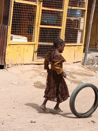 An outdoor scene in Zango, Nigeria, depicts a young female child engaged in play on a dusty, unpaved surface. The child, with dark braided hair, is observed in profile, moving barefoot towards the right side of the frame. She wears a long-sleeved, mustard-yellow top with brown ruffled details on the shoulders, attached to a full, brown, possibly lace-patterned skirt. Her right shoulder is partially exposed. In her right hand, she holds a thin, light-colored wooden stick, positioned to interact with a black rubber tire that rolls upright on the ground to her immediate right.

In the background, a faded yellow building features multiple window openings secured with metal grates. The lower left portion of this building exhibits corrugated metal paneling. Through the grates on the right, a blue container or object is visible inside the structure. The ground beneath the child and tire is uneven, sandy, and scattered with small pieces of white and colored debris. Shadows indicate strong sunlight.