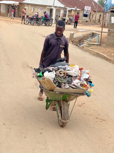 A man in a dark purple shirt with a Yamaha logo is hauling a three-wheeled green cart filled with refuse in Kuchingoro, Nigeria. The cart is overloaded with trash including plastic bags, bottles, and other debris. He is wearing sandals. In the background, there are buildings and people, including people sitting on parked motorcycles. The street is unpaved and dusty.