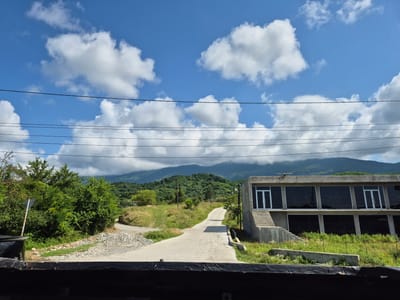 In Adzhiryuara, Abkhazia, a paved road winds its way through a hilly, green landscape. On the right side of the road, a modern, two-story building with large, dark windows stands prominently. The building appears to be under construction or recently completed, with concrete stairs leading up to the entrance. The surrounding area is covered in lush green grass and vegetation. In the background, rolling green hills and mountains are visible under a bright blue sky dotted with white, fluffy clouds. Several power lines are strung across the sky, indicating the presence of infrastructure in the area. The foreground shows the edge of what appears to be a vehicle or covered structure, framing the view of the road and the building. No people are visible in the scene.